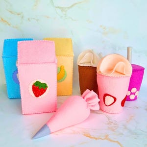 Puede incluir: Una colección de alimentos de fieltro coloridos, que incluyen una manga pastelera rosa con punta plateada y varios recipientes para bebidas. Los recipientes son azules, rosas, amarillos, marrones y morados, con aplicaciones de fresas y plátanos.