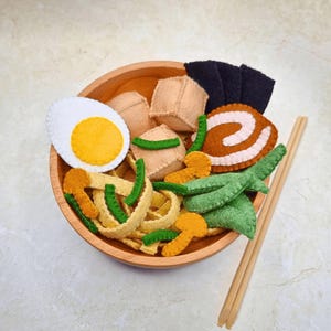 May include: A felt ramen bowl with various toppings, including a boiled egg, tofu, and seaweed. The bowl is made of wood, and chopsticks are placed on the side. The ramen noodles are yellow and green.