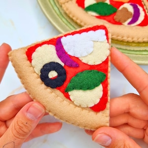 May include: A felt pizza slice with red sauce, white cheese, a black olive, purple onion, and a green leaf. The crust is tan. Another pizza is visible in the background.