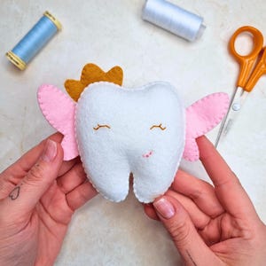 Puede incluir: Un diente de fieltro hecho a mano con alas rosas y una corona dorada. El diente blanco tiene ojos cerrados bordados y una pequeña sonrisa. Suministros de costura, incluyendo hilo y tijeras, están en el fondo.