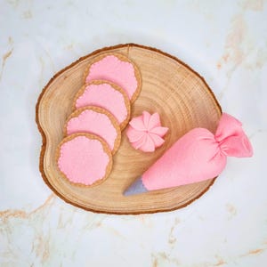 Puede incluir: Una rodaja de madera contiene galletas de fieltro con glaseado rosa, una flor rosa y una manga pastelera rosa con punta gris. Las galletas tienen un borde festoneado. Los artículos están dispuestos sobre una superficie blanca y veteada.