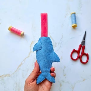 May include: A blue felt dolphin-shaped popsicle holder with a pink popsicle. The holder is hand-stitched and has a detailed design. Also visible are red-handled scissors and spools of thread in pink and blue.