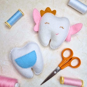 May include: Two white felt tooth-shaped toys, one with a blue pocket and the other with pink wings and a gold crown. The toys are surrounded by sewing supplies, including blue and pink thread spools and orange scissors. The toys have embroidered facial features.