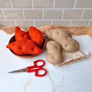 Puede incluir: Un cuenco de madera lleno de batatas de fieltro, junto a patatas de fieltro y unas tijeras rojas. Las patatas son de color beige y las batatas son de color naranja. También hay un pequeño saco de yute.