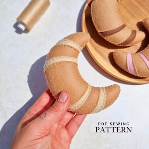May include: A crescent-shaped felt croissant toy in tan with cream-colored stitched stripes. The text PDF SEWING PATTERN is visible in the lower right corner. A spool of tan thread is positioned in the upper left corner near other felt croissants.