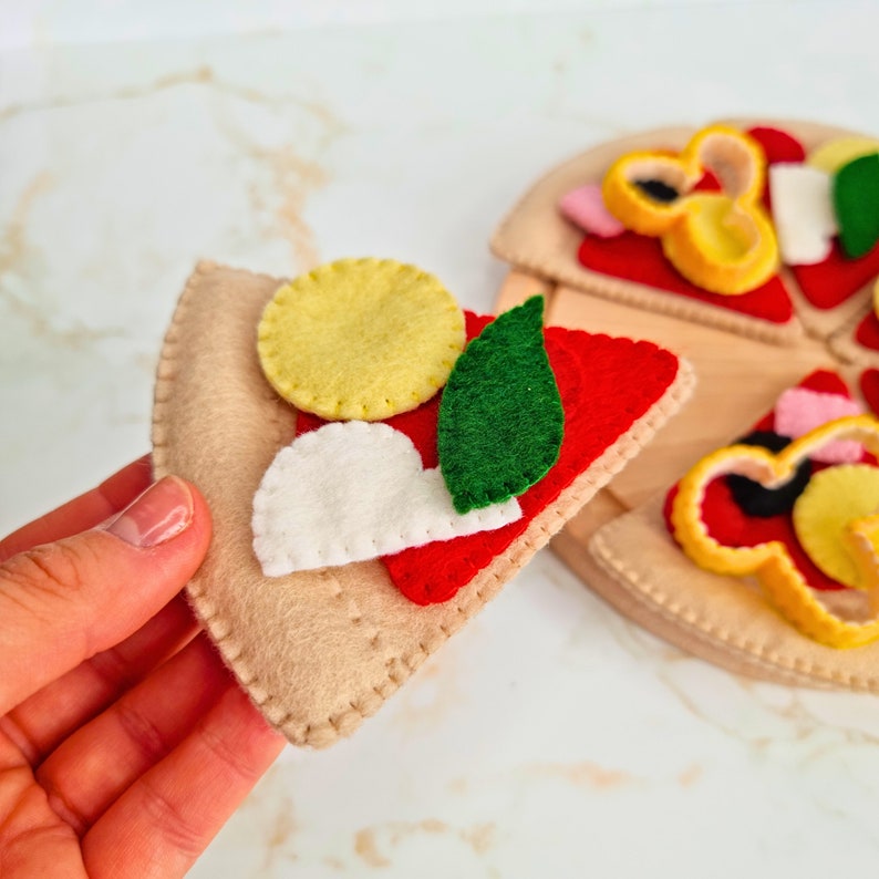 May include: A felt pizza slice toy featuring a tan crust, red sauce, a yellow circular topping, a white semi-circular topping, and a green leaf-shaped topping. The slice is stitched with visible thread along the edges and surface details.
