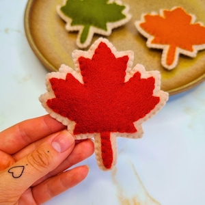 May include: A close-up of a red felt maple leaf with a beige border. Other felt leaves in green and orange are visible in the background. The leaves are on a brown plate. The image is well-lit and shows the details of the felt leaves.