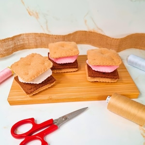 Może przedstawiać: Trzy filcowe s'mores leżą na drewnianej desce. Każdy s'more ma beżowy krakers, białą piankę marshmallow, brązowy kwadrat czekolady i różową piankę marshmallow. Nożyczki z czerwonymi uchwytami i szpulki nici na pierwszym planie.