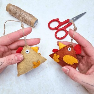 May include: Two felt chicken ornaments in tan and brown, with red and yellow accents. Each has a jute string for hanging. A spool of twine and red-handled scissors are also visible.