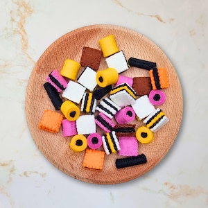 May include: A variety of felt candy shapes in black, white, yellow, pink, and red. The candy shapes are arranged on a wooden plate.