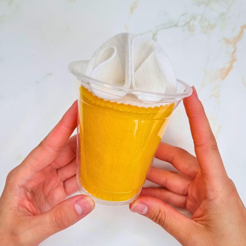 Puede incluir: Un vaso de pl&aacute;stico transparente con un fondo de fieltro amarillo y una parte superior de fieltro blanco que se asemeja a crema batida. El vaso est&aacute; siendo sostenido por dos manos.