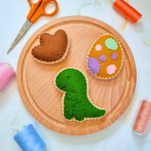 May include: A set of felt appliques featuring a green dinosaur, a brown heart, and a yellow egg with purple and pink polka dots. The appliques are on a wooden surface.