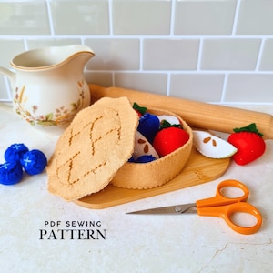 May include: A felt pie crust with a removable lid, filled with plush fruit pieces including red strawberries, blue blueberries, and white apple slices. The set includes orange scissors and the text PDF SEWING PATTERN printed on the surface.