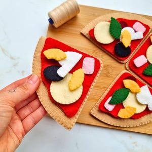 May include: Felt pizza slices featuring a tan crust, red sauce, and various toppings including yellow, white, black, and pink shapes. The set includes multiple triangular pieces arranged on a wooden board with a spool of tan thread nearby.