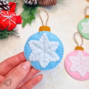 May include: A round, light blue felt Christmas ornament featuring a white stitched snowflake design in the center and a mustard yellow felt cap at the top. The ornament includes a thin brown twine loop for hanging, with visible stitching along the edges.