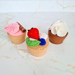 Può includere: Tre cupcake di feltro con diverse guarnizioni. Un cupcake ha glassa rosa e un bastoncino di cioccolato, un altro ha una fragola, un kiwi e un mirtillo, e l'ultimo ha glassa bianca.