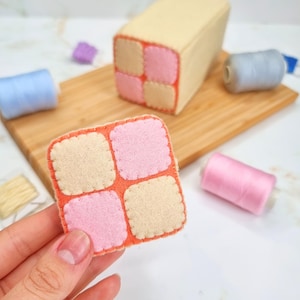 May include: A felt patchwork square with four squares, two pink and two beige, sewn together with orange thread. The squares have a scalloped edge.