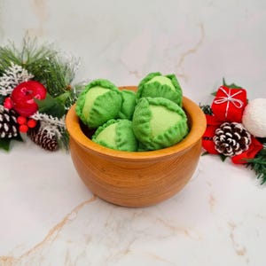 Op de afbeelding: Een houten kom gevuld met vilt spruitjes. De kom staat op een wit marmeren oppervlak. Er zijn kerstversieringen op de achtergrond.