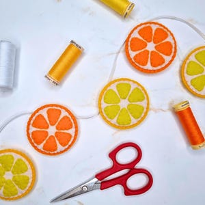 May include: A handmade felt garland featuring orange and lemon slices, strung on a white cord. The garland is accompanied by spools of yellow, orange, and white thread, and a pair of red-handled scissors. The design is bright and cheerful.