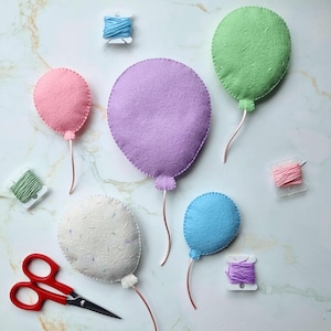 Pode incluir: Balões de feltro feitos à mão em tons pastel: rosa, roxo, verde, azul e branco. Cada balão tem um caule de arame de cobre. Também estão presentes carretéis de linha e tesouras vermelhas. Imagem com tema artesanal.