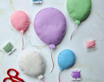 May include: Five felt balloon shapes in pink, purple, light green, white with colorful flecks, and blue. Each balloon has a thin string attached. Included are four small spools of embroidery thread in blue, pink, green, and purple, and red scissors.