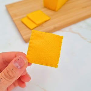 Puede incluir: Una rebanada de queso cuadrada de fieltro amarillo brillante se sostiene en la mano. Dos rebanadas adicionales y un bloque de queso están sobre una tabla de cortar de madera. Las rebanadas de queso son de un color vibrante y sólido.
