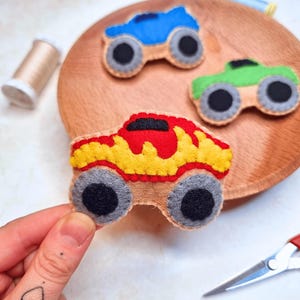 Puede incluir: Tres juguetes de fieltro con forma de monster truck de colores. Uno es azul, otro verde y otro rojo y amarillo. Cada camión tiene ruedas grises con centros negros. Los juguetes están sobre un plato de madera.