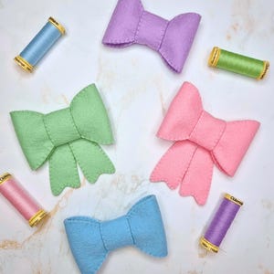 May include: An assortment of colorful felt bows in pastel shades of pink, green, blue, and purple. The bows are arranged with spools of thread in matching colors, creating a craft-themed composition.