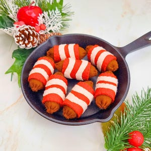 Puede incluir: Una sartén de hierro fundido negro llena de siete adornos de fieltro con forma de salchicha envueltos en rayas rojas y blancas. Los adornos son marrones en los extremos. Hay vegetación decorativa y una manzana roja en el fondo.