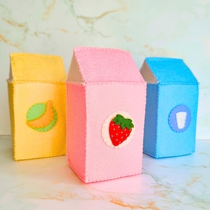 May include: Three colorful felt milk carton-shaped containers. One is yellow with a banana, one is pink with a strawberry, and the last is blue with a glass of milk. The containers have a stitched design and a white top.