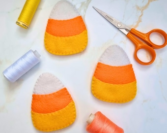 May include: Three felt candy corn shapes featuring horizontal stripes of white, orange, and yellow. The set includes a spool of yellow thread, a spool of white thread, a spool of orange thread, and a pair of orange-handled metal scissors.