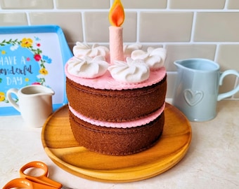 May include: A two-tiered brown felt cake with a pink felt top layer, white felt dollops, and a single pink felt candle with an orange and yellow flame. The cake sits on a round wooden plate, accompanied by orange-handled scissors.