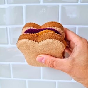 Puede incluir: Un juguete de sándwich de fieltro que consta de dos rebanadas de pan de color tostado claro, una capa de relleno amarillo y una capa de relleno morado. Las piezas están unidas con costuras beige visibles a lo largo de los bordes, creando un objeto.