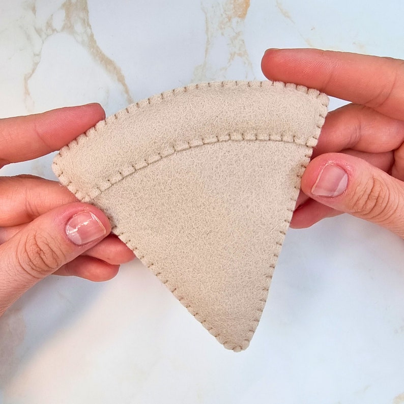 May include: A triangular piece of beige felt fabric featuring a rounded top edge and visible whip stitching along the perimeter. The felt material has a soft, matte texture and is constructed with a distinct seam separating the crust area.