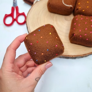 以下が含まれることがあります： 手で持たれた、カラフルな刺繍ドットが付いた茶色のフェルト正方形。他の同様の正方形が木の輪切りにあります。背景には赤いハサミがあります。正方形はミニチュアブラウニーに似ています。
