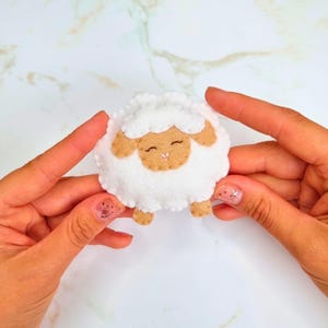 Puede incluir: Un pequeño adorno circular de fieltro con forma de oveja. La oveja tiene un cuerpo de lana blanca texturizada, una cara de color tostado con los ojos cerrados y una pequeña nariz cosida. Se ven dos orejas y dos pequeños pies tostados.