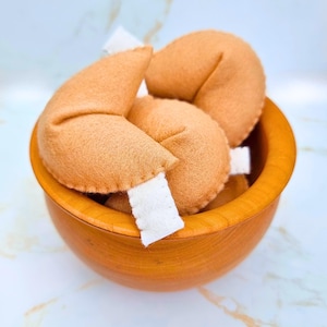 Puede incluir: Un cuenco de madera lleno de varias galletas de la fortuna de color marrón claro. Cada galleta tiene una tira de papel blanco asomando. El cuenco es de madera marrón claro y está sobre una superficie blanca y azul claro.