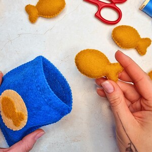 May include: A bright blue felt pouch featuring a circular tan patch with a gold fish silhouette. Several small, gold, fish-shaped felt cutouts are scattered nearby, alongside red scissors and a spool of blue thread for crafting projects.
