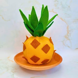 May include: A felt pineapple with green leaves and brown diamond-shaped details, presented on a small wooden plate. The pineapple is yellow and has a textured finish. Ideal for kitchen or tropical-themed decor, this item is handmade.