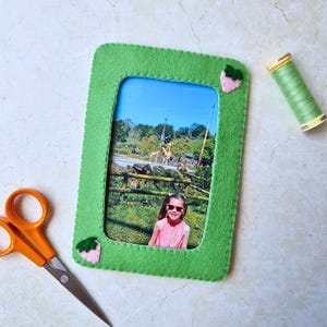 Puede incluir: Un marco de fotos rectangular de fieltro verde con bordes cosidos y dos pequeñas aplicaciones de fresas rosas y verdes. El marco contiene una fotografía de un niño con gafas de sol rosas frente a un recinto de jirafas en un zoológico.