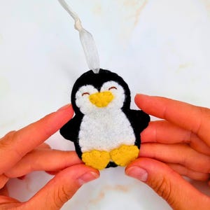 May include: A handmade felt penguin ornament with a white belly, black body, and yellow beak and feet. The penguin has a smiling face and a white ribbon for hanging. A cute, cheerful holiday decoration.