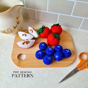 Peut inclure: Une collection de fruits en feutrine comprenant trois fraises rouges à feuilles vertes, sept myrtilles bleues avec des détails cousus et deux tranches de pomme blanches avec des pépins marron, sur une planche à découper en bois avec des ciseaux orange.