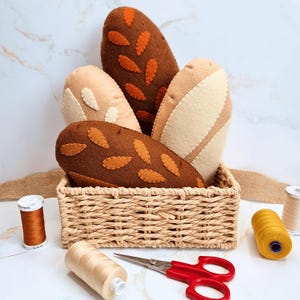 May include: A woven basket filled with plush bread loaves in various shades of brown and beige. The loaves have decorative felt accents. Sewing supplies, including thread spools and red scissors, are arranged around the basket.