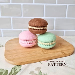 May include: Three felt macaron replicas in brown, pink, and light green, each featuring a cream-colored stitched filling between two rounded shells. The text PDF SEWING PATTERN is printed in the lower right corner of the image.