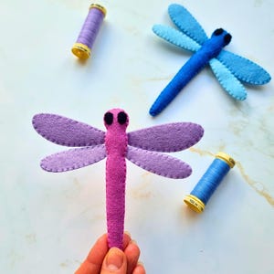 May include: Handmade felt dragonflies in shades of purple, pink, and blue. The dragonflies have black eyes and are stitched with contrasting thread. Two spools of thread in purple and blue are also in the image, adding a craft-like aesthetic.