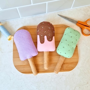 Puede incluir: Tres juguetes de paletas de fieltro con detalles cosidos. Una es de color lavanda sólido, otra es rosa con una cobertura de chocolate marrón y chispas de colores, y otra es verde claro con pequeños detalles cosidos en marrón.