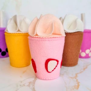Puede incluir: Cinco vasos de juguete de fieltro con tapas de crema batida y bebidas de diferentes colores. Los vasos son amarillos, rosas, marrones, morados y rosas. El vaso rosa tiene diseños de fresas.