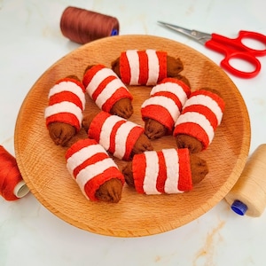 Puede incluir: Un plato de madera contiene varias creaciones de fieltro hechas a mano que se asemejan a artículos envueltos en tocino. Cada pieza es marrón con rayas rojas y blancas alternas. También se ven carretes de hilo y tijeras rojas.