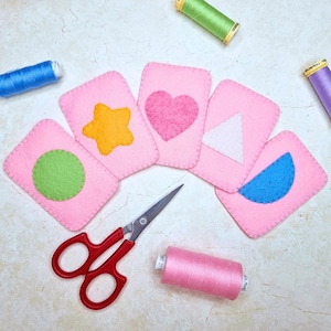 Könnte beinhalten: Rosa Filzkarten mit verschiedenen Formen: ein grüner Kreis, ein gelber Stern, ein rosa Herz, ein weißes Dreieck und ein blauer Halbmond. Rote Schere und Garnrollen in Rosa, Grün, Blau und Lila.