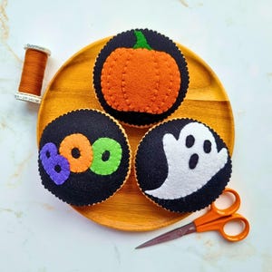 May include: Three circular felt ornaments featuring Halloween designs: an orange pumpkin, the word BOO in purple, orange, and green, and a white ghost on a black background. Includes a spool of orange thread and orange-handled scissors.
